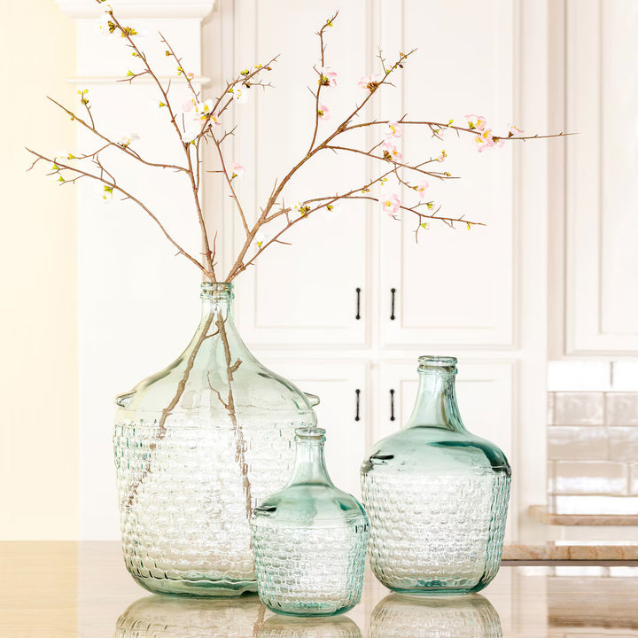 Three textured glass vases with branches on a reflective surface