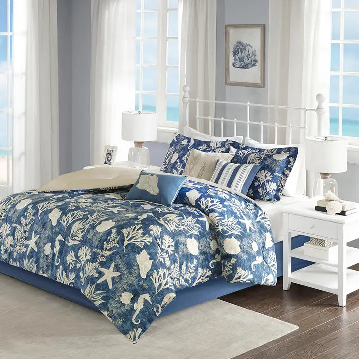 A navy blue comforter set with a floral pattern of seashells displayed on a bed in a room with a white headboard and pillows, alongside a bed skirt and decorative pillows.