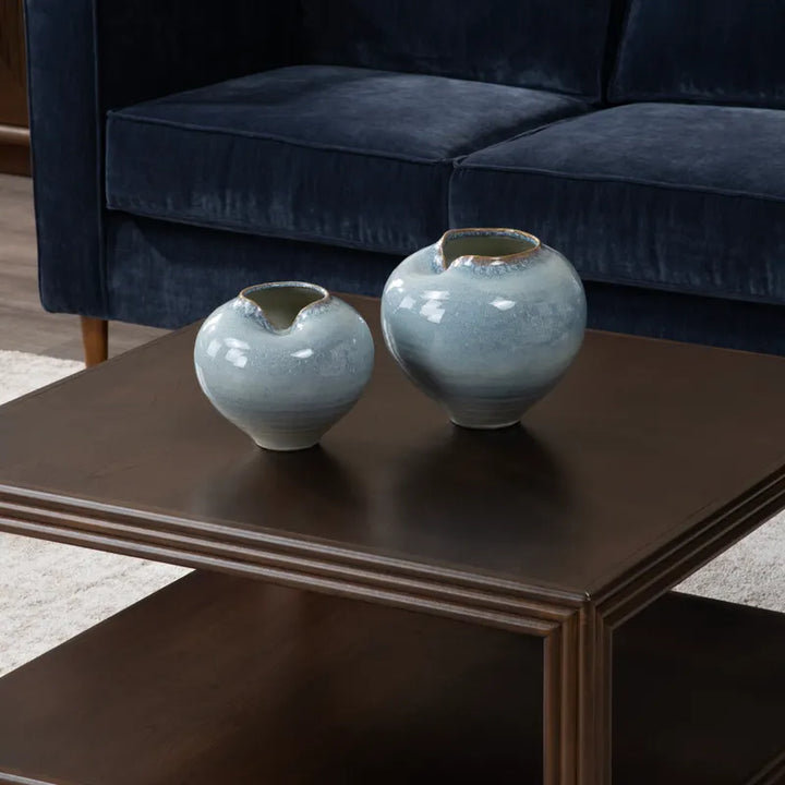 Two light blue-green ceramic vases with a glossy glaze and small slits for pouring, displayed on a dark wood coffee table in front of a navy blue velvet sofa.