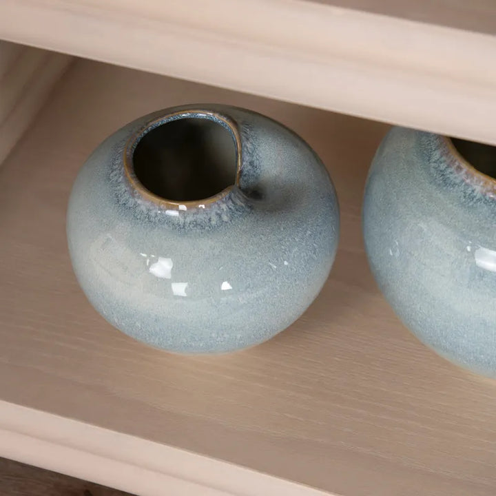 A small round ceramic vase with a glossy light blue and white speckled glaze, featuring a unique asymmetrical slant opening, displayed on a light wood shelf next to another similar vase.