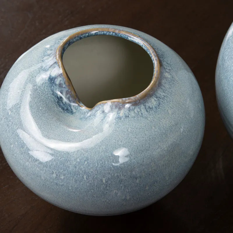A close-up of a round ceramic vase with a glossy speckled light blue-gray glaze and a rustic irregular rim, shown on a dark wooden surface.