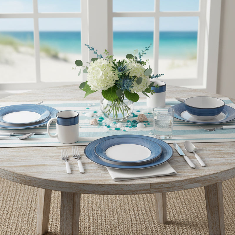Dining table set with blue and white tableware, flowers, and glasses by a window with a beach view.