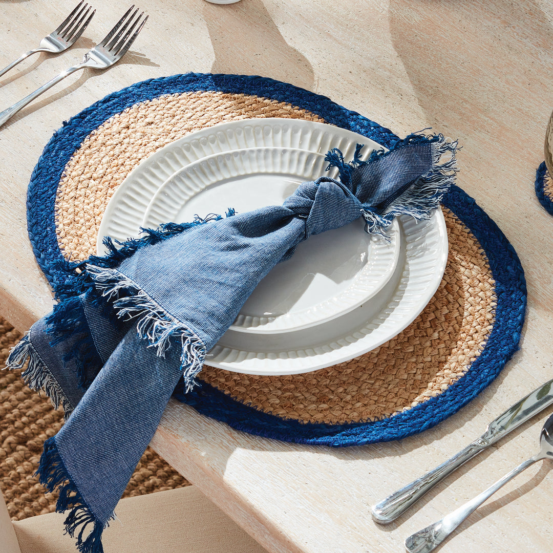 Table setting with blue napkin, white plates, and jute placemats with blue border on a wood table.