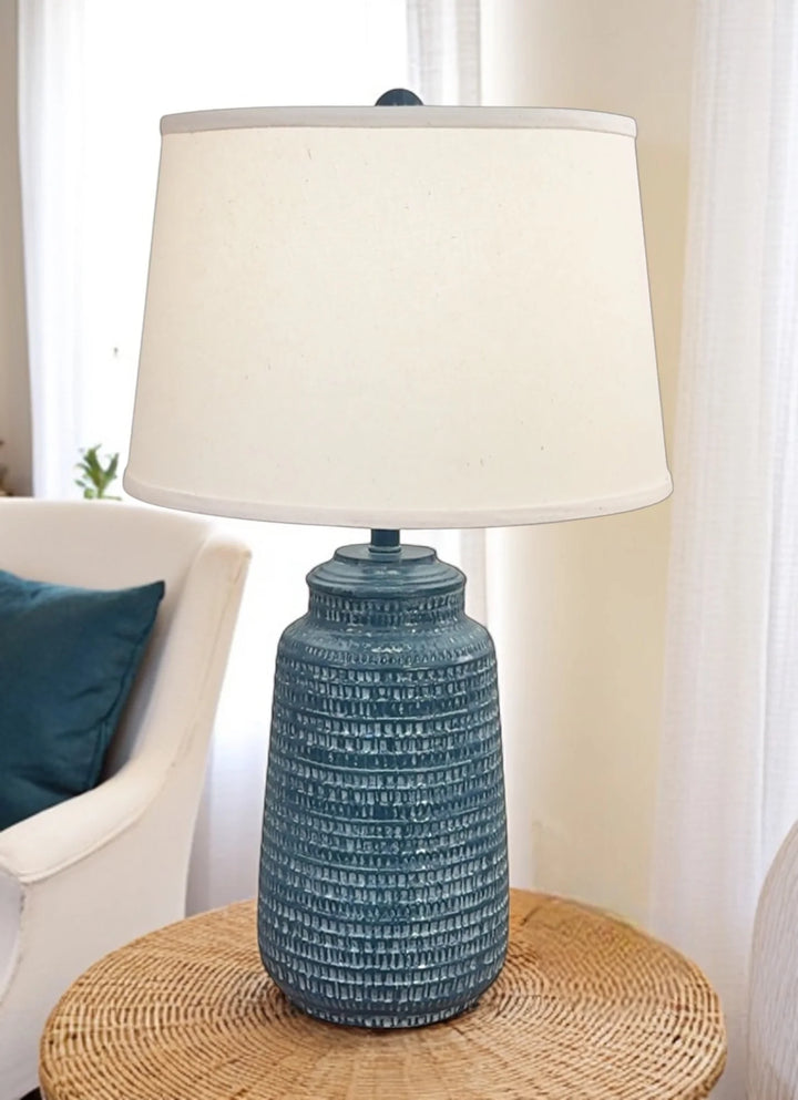 Blue textured lamp with a white shade on a wicker side table in a living room setting.