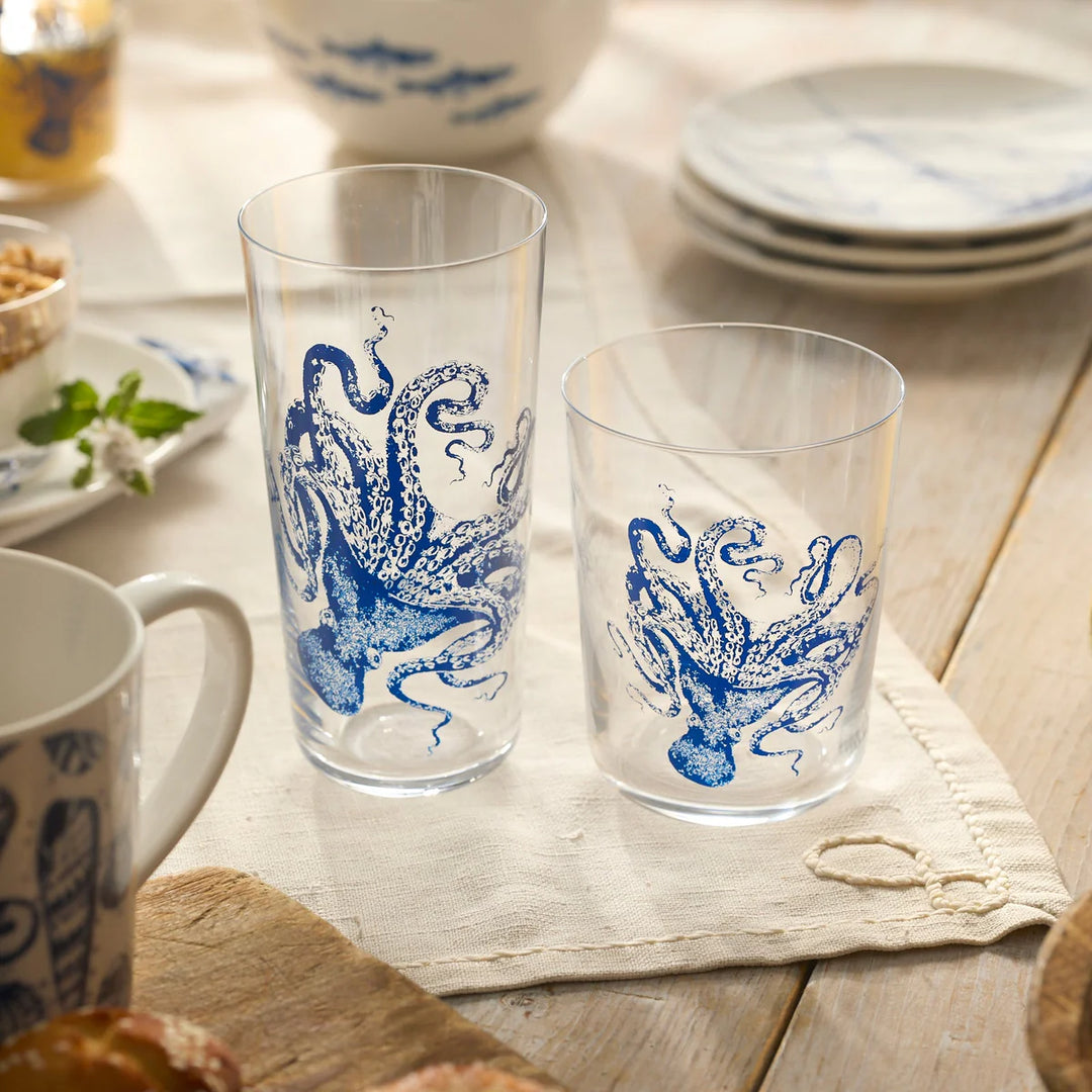 Two glass mugs with blue octopus designs on a wooden table with plates and bowls in the background.