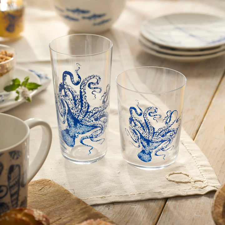 Two glass mugs with blue octopus designs on a wooden table with plates and bowls in the background.