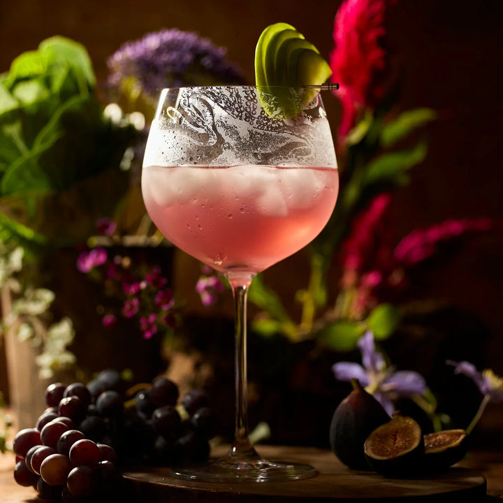 Pink cocktail in a glass with a decorative garnish, surrounded by grapes and figs on a wooden surface. Coastal Compass Home Decor
