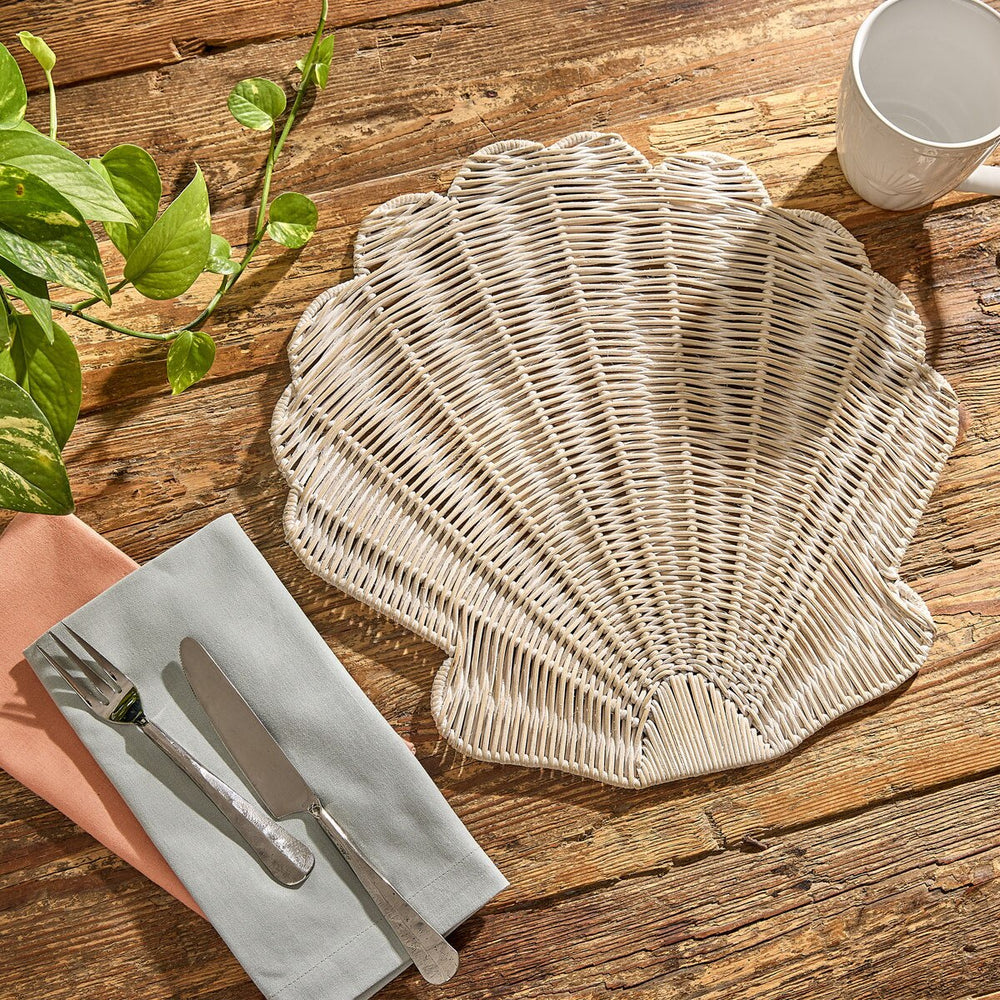 Woven scallop seashell placemat on a wooden table with cutlery and a cup.