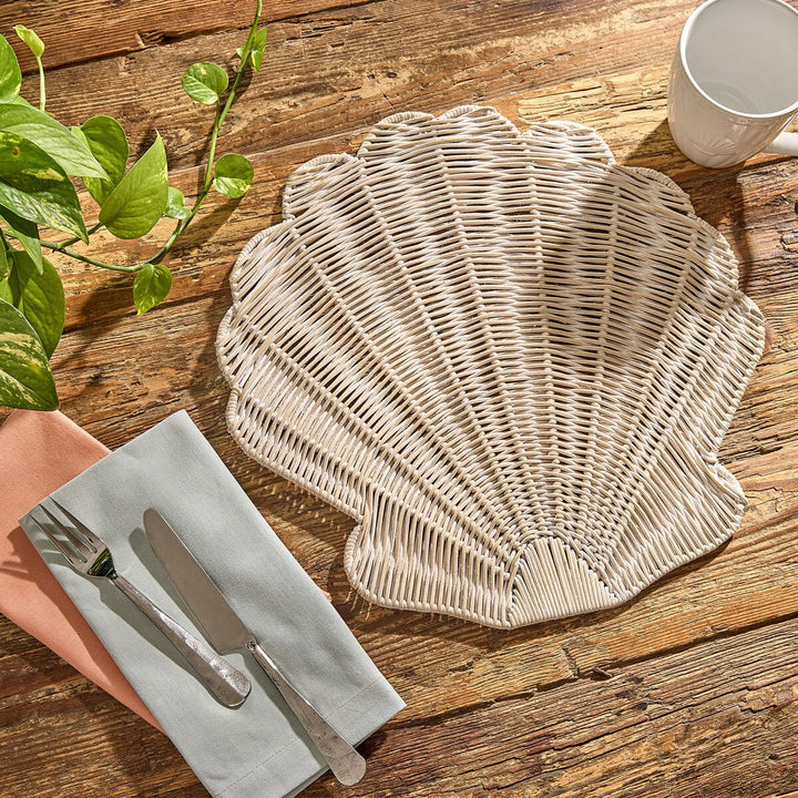 Woven scallop seashell placemat on a wooden table with cutlery and a cup.