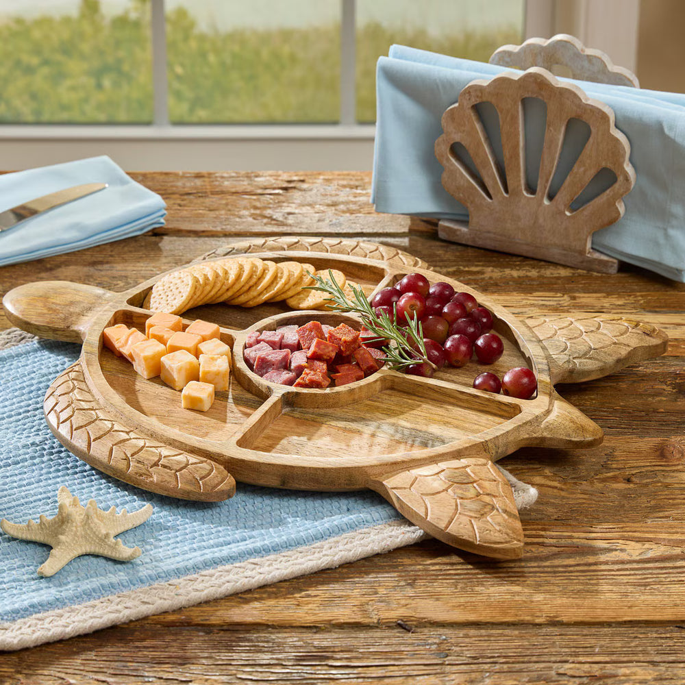 A carved wooden turtle-shaped charcuterie board with five compartments, styled on a rustic wooden table with crackers, cheese, grapes, and a napkin holder in the background.