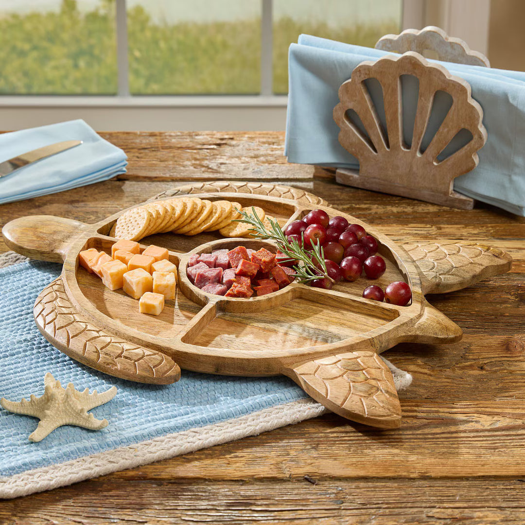 A carved wooden turtle-shaped charcuterie board with five compartments, styled on a rustic wooden table with crackers, cheese, grapes, and a napkin holder in the background.