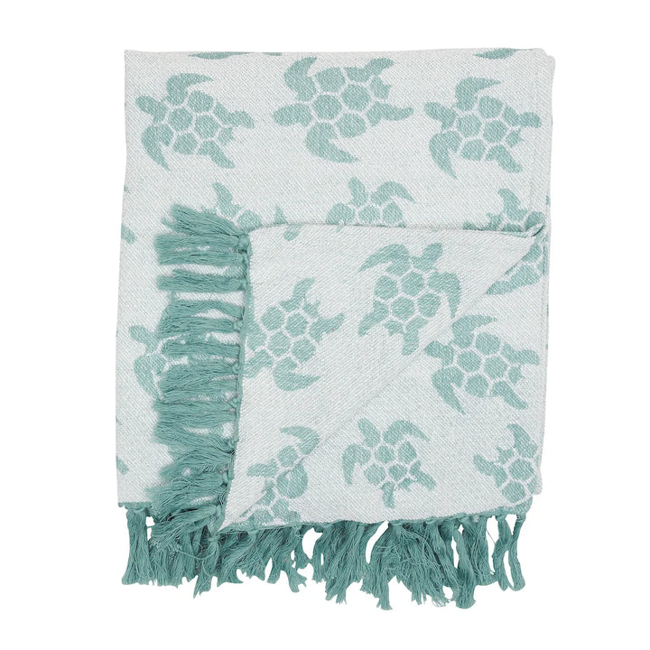 A folded woven throw blanket with a sea turtle pattern in a muted teal-green on a white background, featuring fringed edges.