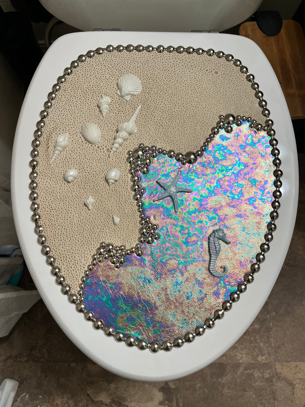 Decorative Sea & Shore toilet seat with shells, starfish, and seahorse design on a beige leather background.