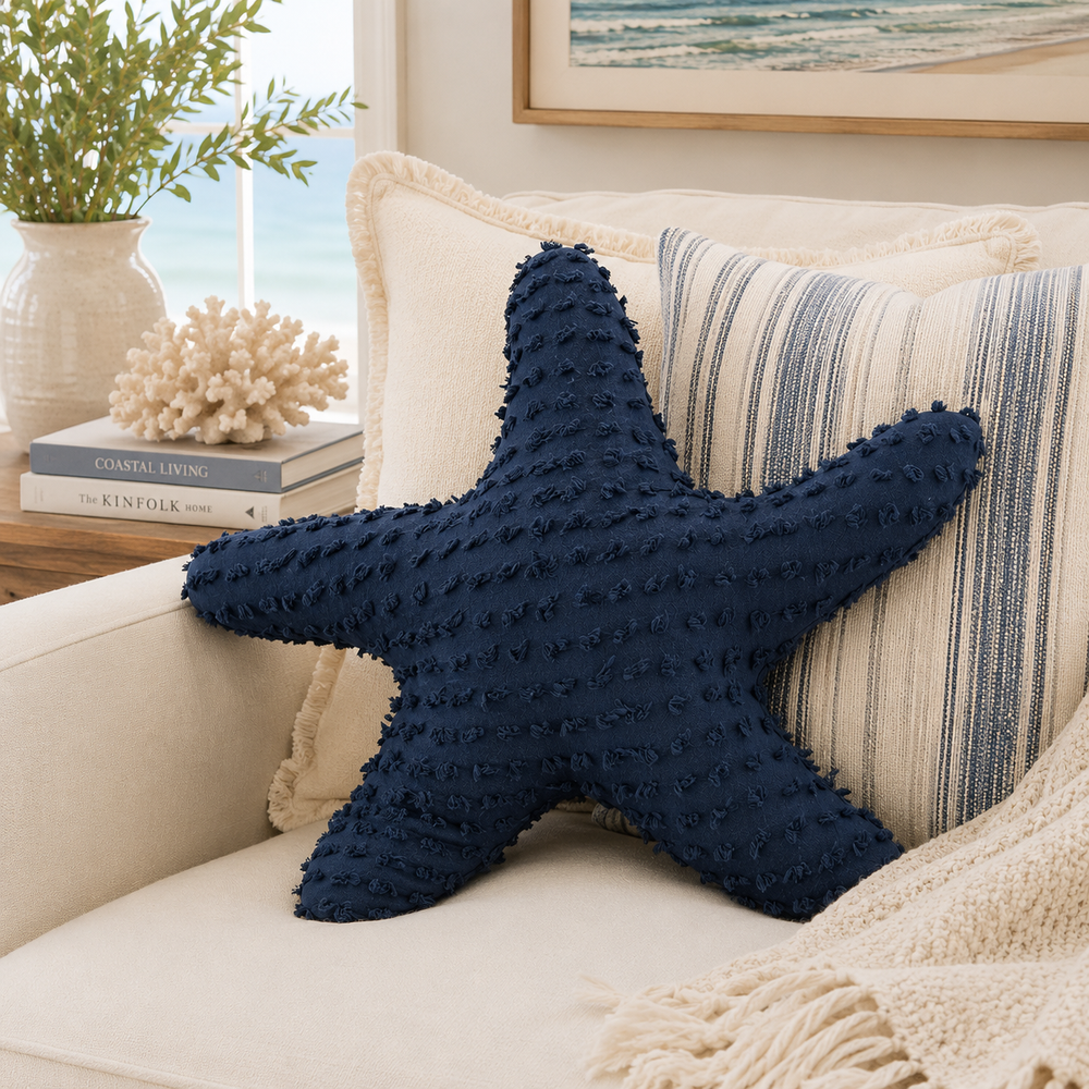 A navy blue starfish-shaped textured throw pillow resting on a cream-colored sofa next to striped pillows and a knit blanket.