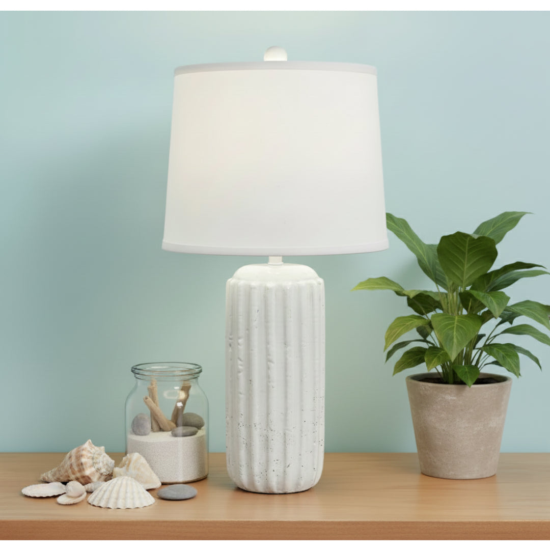 White textured table lamp on a wooden surface with a plant and decorative items against a light blue wall.