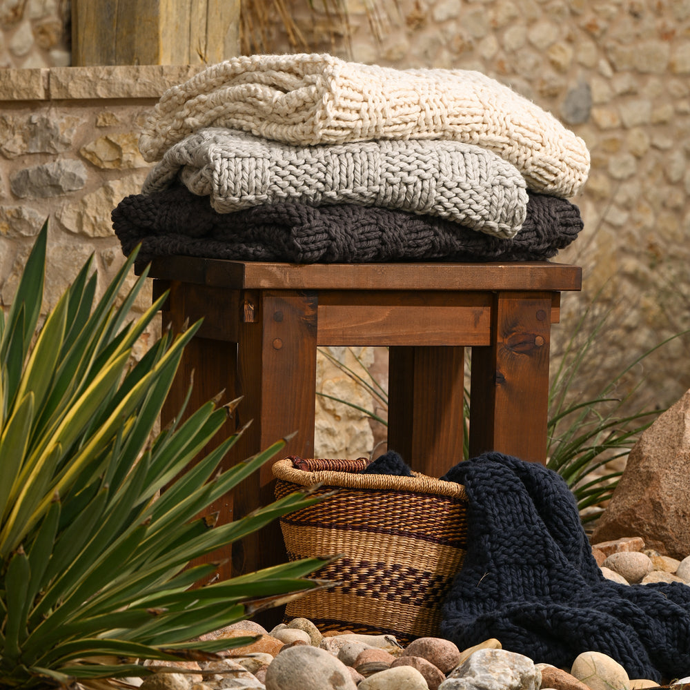Stack of knitted blankets on a wooden stool with a stone wall and plants in the background - Coastal Compass Home Decor