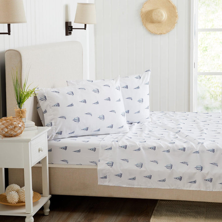 Bed with white bedding featuring blue sailboat pattern, nightstand with decorative items, and lamp in a bedroom setting.