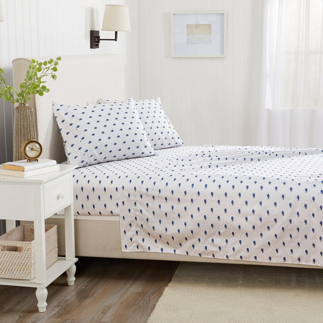 Bedroom with a bed featuring a white comforter with blue patterns, a nightstand with books and a plant, and a lamp on a white wall.