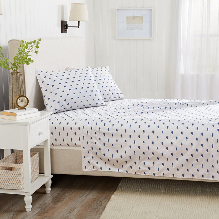 Bedroom with a bed featuring a white comforter with blue patterns, a nightstand with books and a plant, and a lamp on a white wall.