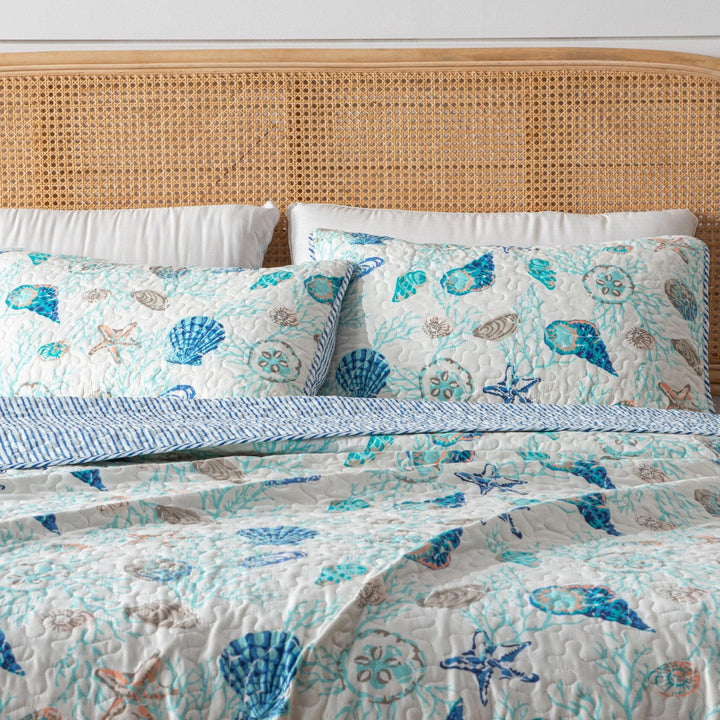 Bed with a coastal shells patterned quilt and pillows against a rattan headboard.