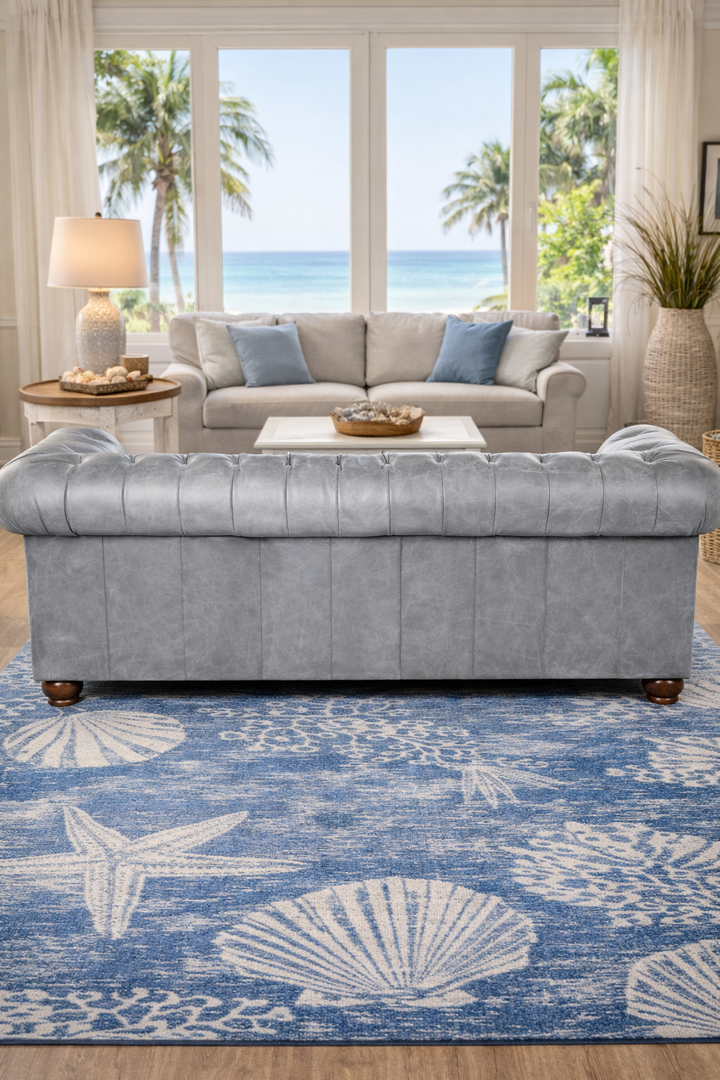 Coastal living room showing the back of the Harbor Fog grey leather Chesterfield Sofa on blue and white sea shell coastal rug with ocean view 