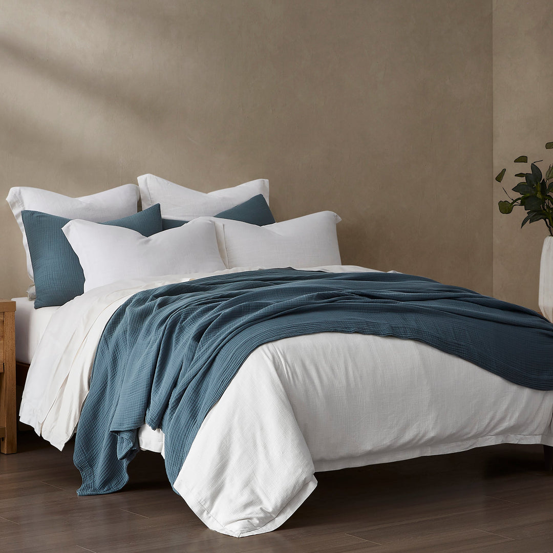 Bed with white bedding and voile cotton storm blue blanket set against a beige wall
